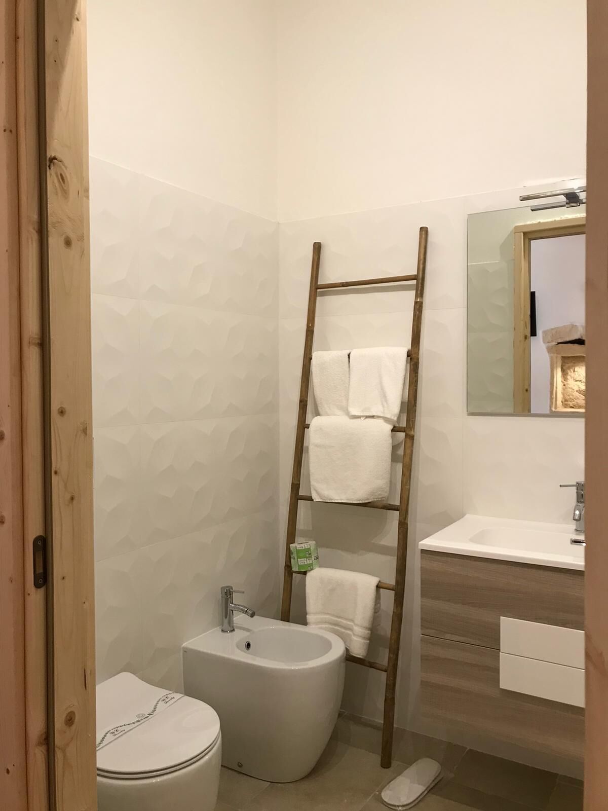 A bathroom with a toilet, bidet, wooden ladder with towels, and a mirror above a sink.