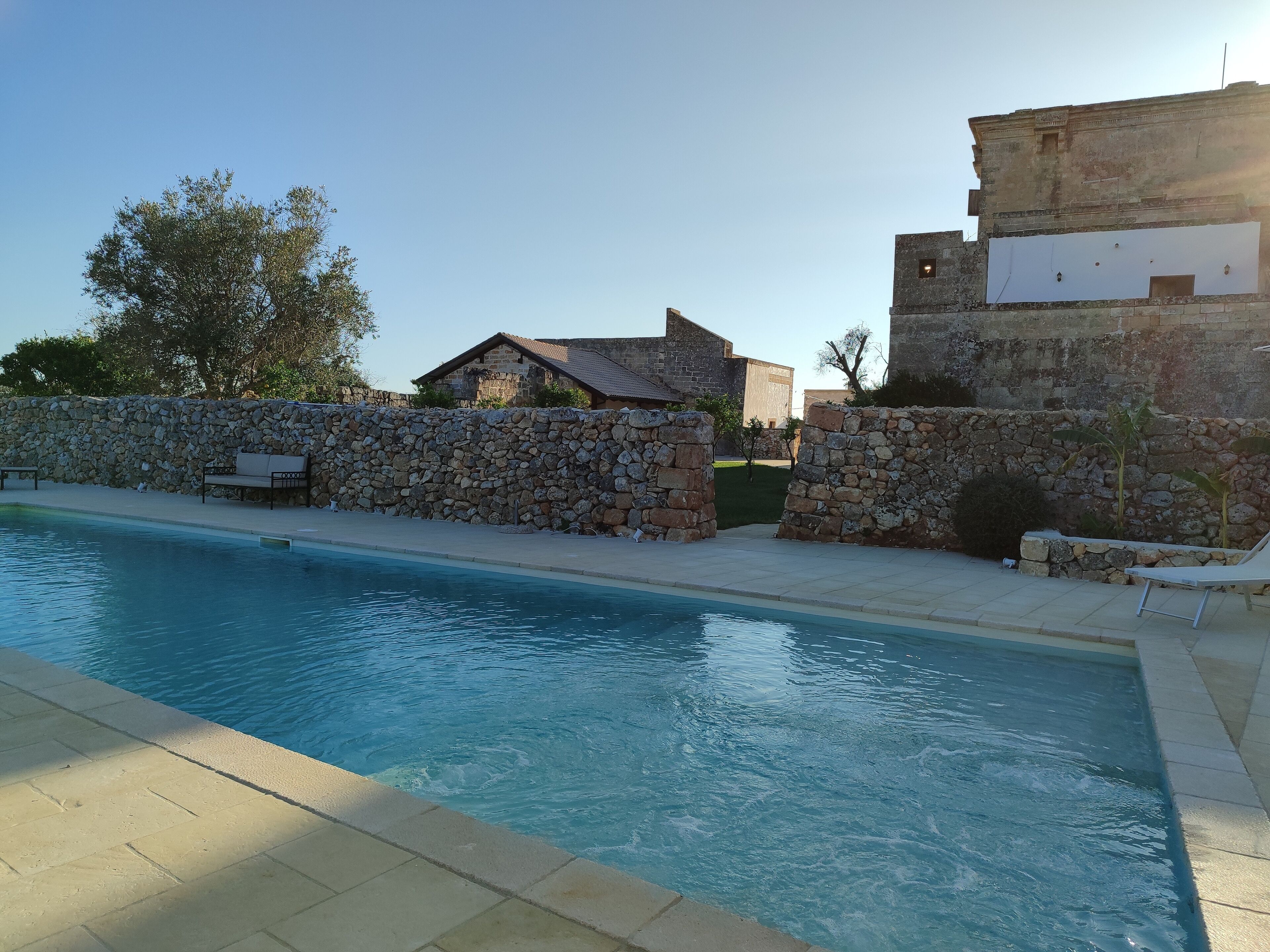 Una piscina con paredes de piedra y un banco, rodeada por un muro de piedra y un edificio con tejado de baldosas.