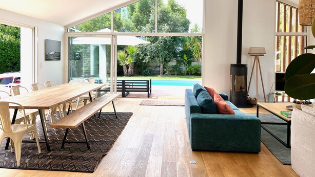 Een ruime woonkamer met een houten eettafel, een groene bank en uitzicht op een zwembad en tuin.