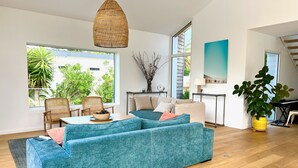 Een ruime woonkamer met een grote blauwe bank, een rieten hanglamp en uitzicht op groen.
