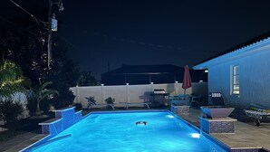 Ein rechteckiger Swimmingpool mit Fliesenrand, umgeben von einer Terrasse mit Gartenmöbeln und einer überdachten Veranda.