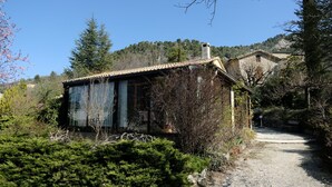 A rustic stone building with a tiled roof, surrounded by dense greenery and a clear sky.