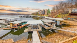 En småbåtshamn vid vattnet med förtöjda båtar, en träbrygga och bostadshus i bakgrunden.