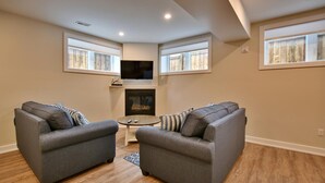 A living room with two grey sofas, a small round table, a fireplace, and a TV mounted on the wall. - The Landing - Extended Stay Quarters (Meaford)