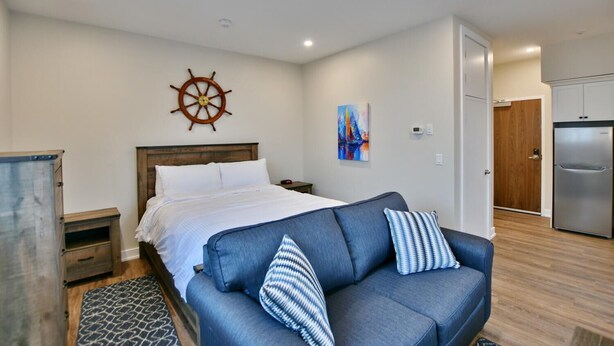 A bedroom with a bed, a wooden headboard, a ship's wheel decoration, a painting, a blue sofa with patterned cushions, a small table, and a refrigerator in the adjacent room. - The Landing - Extended Stay Quarters (Meaford)