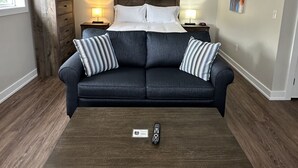 A living room with a dark blue sofa, two pillows, a wooden coffee table, a ship's wheel wall decoration, a lamp, and a painting on the wall. - The Landing - Extended Stay Quarters (Meaford)
