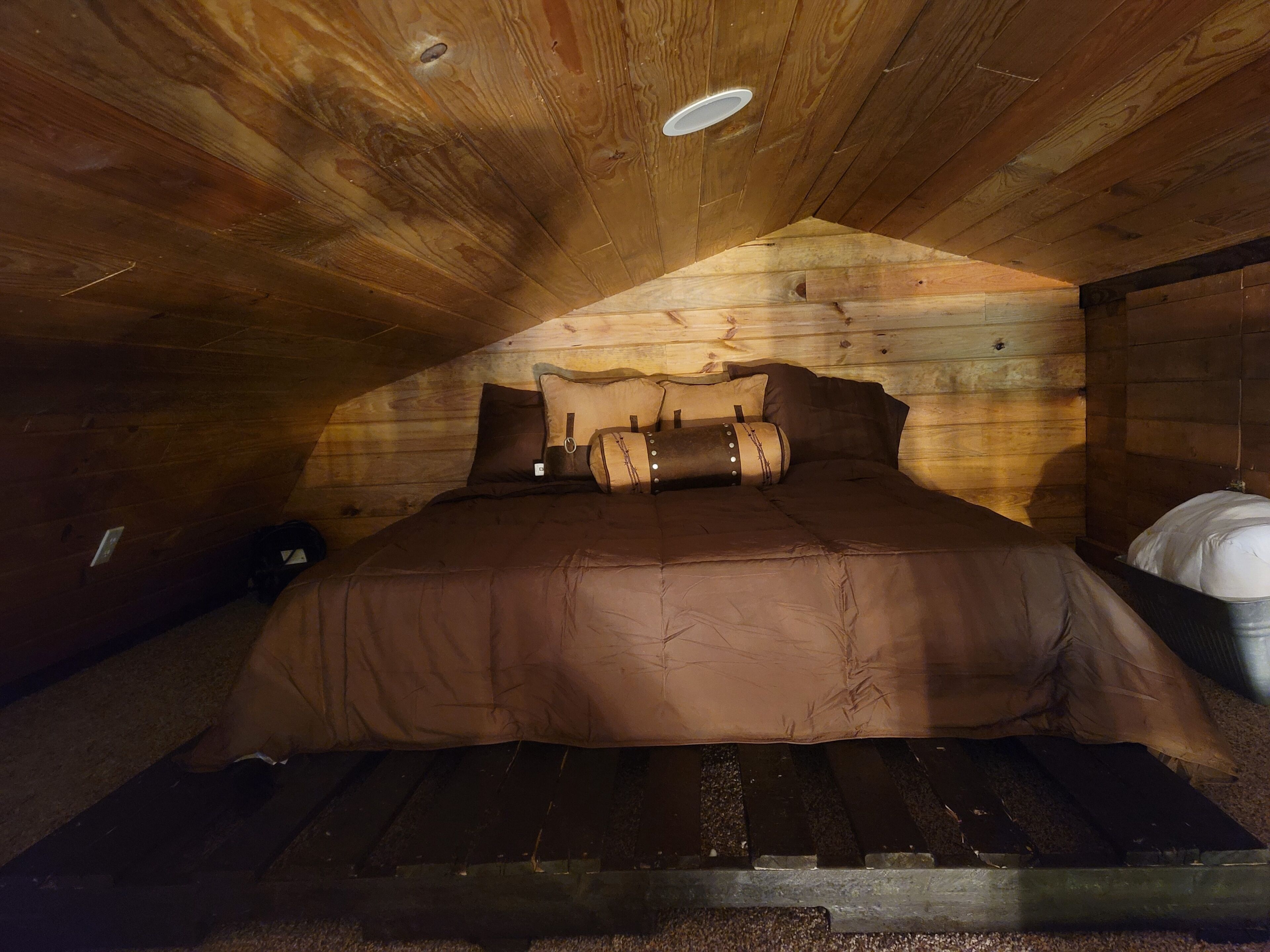Une chambre mansardée chaleureuse avec un plafond en pente en bois, un lit avec une literie marron et une petite table ronde.