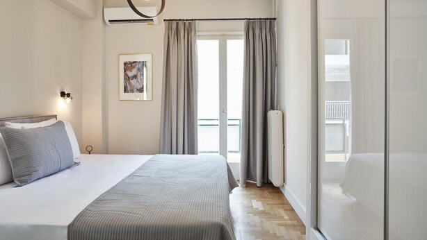 A bedroom with a bed, a bedside table, a wall-mounted light, a window with curtains, and a radiator. - Ilisia Park Contemporary 2BD Condo (Athens)