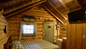 Une chambre confortable aménagée dans un chalet en rondins, avec un lit, une commode, une fenêtre avec des rideaux et un téléviseur fixé au mur.