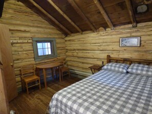 Una habitación de cabaña de madera con una cama, una mesita pequeña, dos sillas, un ventanal y un cuadro en la pared.
