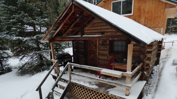 Une cabane en bois dotée d’un porche abrité, d’une échelle menant à l’entrée et d’un toit recouvert de neige.