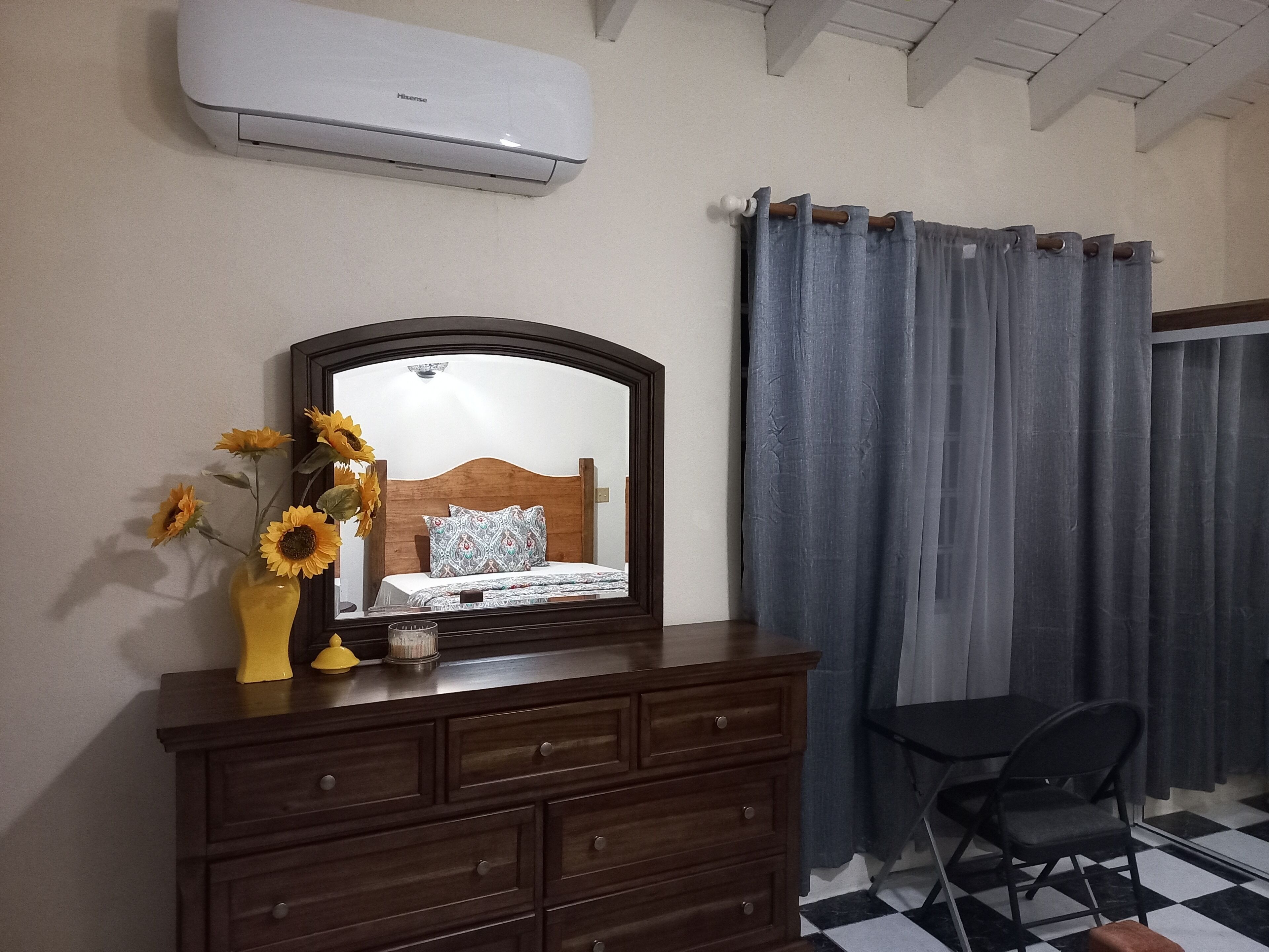 Um quarto com cômoda de madeira, um espelho, uma cama com roupa de cama estampada, um vaso com girassóis e uma janela com cortinas cinzas.
