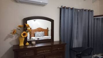 Um quarto com cômoda de madeira, um espelho, uma cama com roupa de cama estampada, um vaso com girassóis e uma janela com cortinas cinzas.