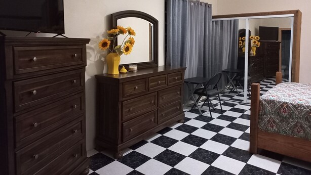 Un dormitorio con suelo a cuadros blanco y negro, un aparador de madera, un espejo, un televisor, una cama con ropa de cama estampada, un ventilador de techo y un equipo de aire acondicionado.