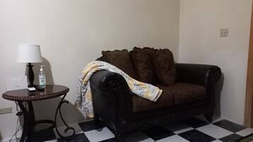 A living room with a checkered floor, a dark brown sofa, a small round table with a lamp, and a bottle of hand sanitizer.