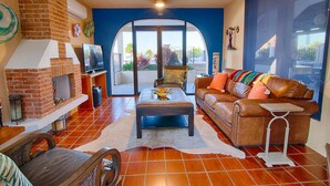 Un salon doté d’une cheminée en briques, d’un canapé en cuir, d’une table basse et offrant une vue sur un balcon extérieur, accessible par une porte vitrée.