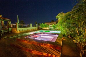 Una piscina con jacuzzi, rodeada de palmeras y una cerca de noche.