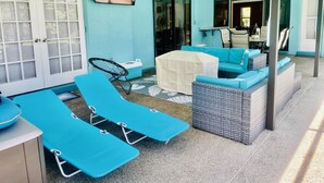 A sunlit room with blue walls, a ceiling fan, a flat-screen TV, a covered outdoor seating area, and a view into the adjacent room.