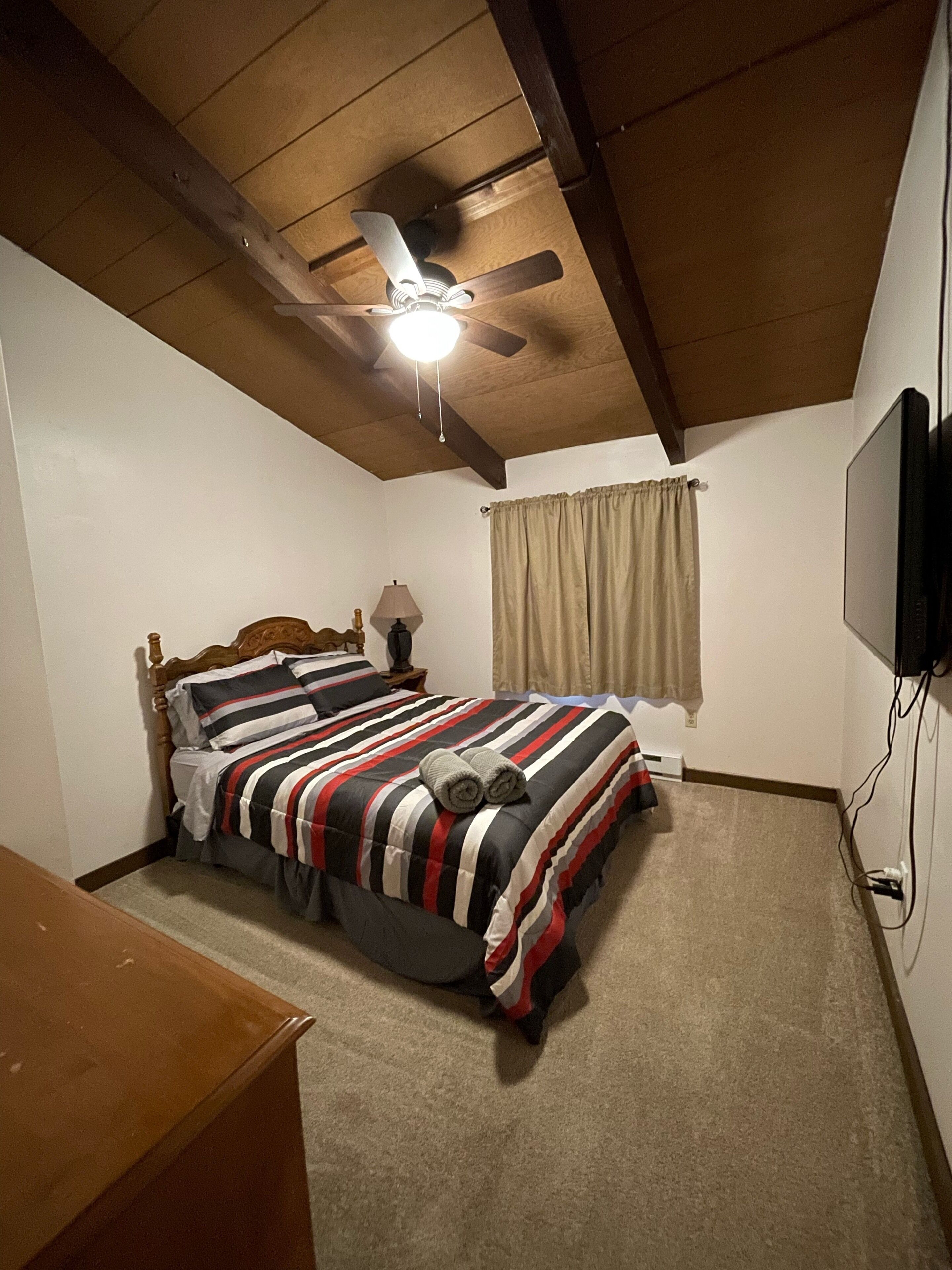 Un dormitorio con cama, ventilador de techo, lámpara, televisor y ventana con cortinas.