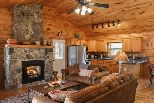 A cozy log cabin interior with a stone fireplace, leather sofa, and a kitchen area.