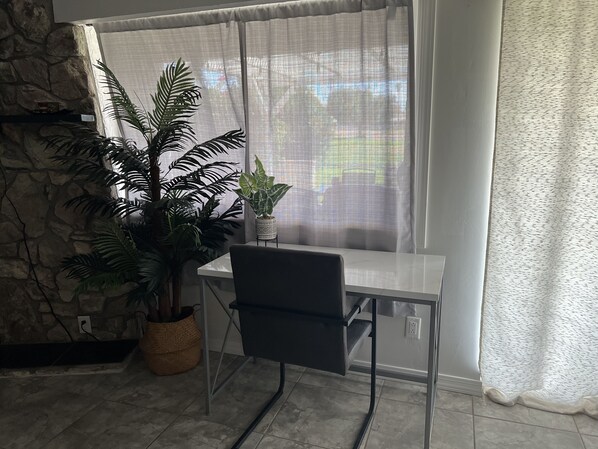 Una habitación con pared de piedra, una ventana con cortinas traslúcidas, una planta en maceta, un escritorio con una silla y suelo de baldosas.
