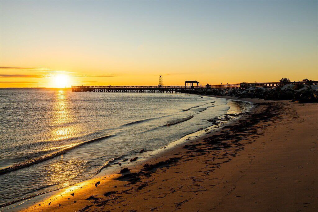 Ein Strand mit einem Steg, der sich ins Wasser erstreckt, bewölkter Himmel und Sonnenauf- oder -untergang.