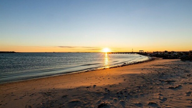 Una playa al atardecer con un mar tranquilo, una orilla arenosa y un muelle que se adentra en el agua.