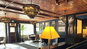 Um hall com um lustre imponente, uma mesa grande com abajur amarelo e quadros nas paredes.