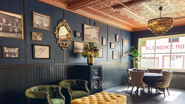 Um quarto com parede revestida de madeira escura, um candelabro, quadros emoldurados, um sofá verde, um banquinho amarelo e um espaço de jantar com cadeiras e uma mesa.