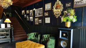 Um quarto com escada, poltronas estofadas em verde, um banquinho dourado com capitonê, um rádio antigo e uma parede decorada com fotografias emolduradas.