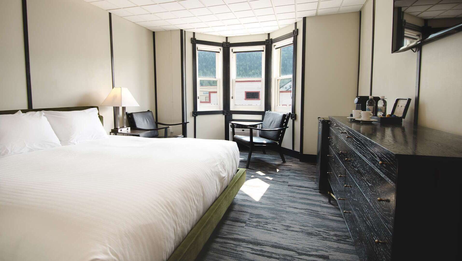 Ein Hotelzimmer mit einem großen Bett, einem Schreibtisch mit Stuhl, einem Schminktisch und einem Fenster mit Blick auf Gebäude.