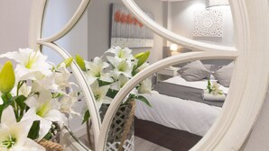 Een ronde spiegel met een witte lijst aan een muur, die een slaapkamer weerspiegelt met een bed, kussens en een nachtkastje.