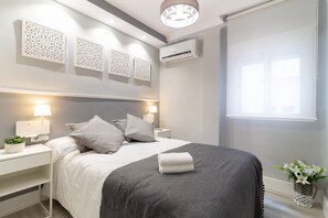 Ein modernes Schlafzimmer mit einem ordentlich bezogenen Bett, Nachttischen mit Lampen, Wandkunst und einer Klimaanlage. Ein Fenster sorgt für natürliches Licht.