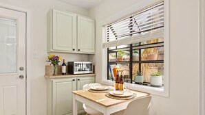 Une cuisine avec des meubles blancs, une table en bois et une fenêtre avec des stores.
