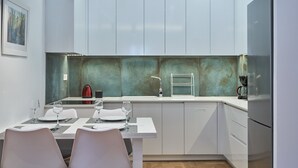 Une cuisine moderne avec des meubles blancs, un réfrigérateur en acier inoxydable et une table à manger pour deux personnes, agrémentée de verres à vin et d’assiettes.