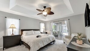 Une chambre à coucher comprenant un lit, des tables de chevet, un ventilateur de plafond, un bureau avec une plante et une porte-fenêtre coulissante donnant sur un balcon.