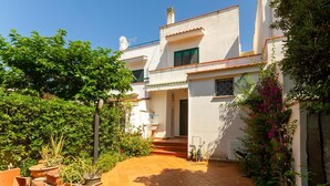 Una casa de dues plantes amb un pati enrajolat, un petit jardí amb plantes en testos i una porta que dona a l'interior.