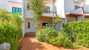 Un pati amb un camí de maó, envoltat de vegetació abundant i un edifici blanc amb persianes verdes.