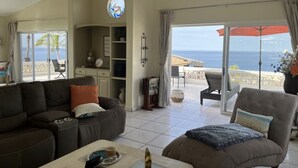 A living room with a sofa, armchair, and coffee table, a ceiling fan, and a view of the ocean through large sliding doors. - Beautiful Ocean View Home (Cabo San Lucas)