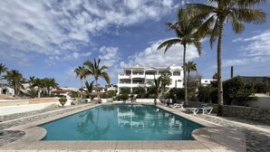 A swimming pool with a stone border, surrounded by palm trees and a white building with balconies. - Beautiful Ocean View Home (Cabo San Lucas)