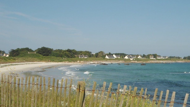 A coastal beach scene with a wooden fence in the foreground, a sandy beach, and houses in the background.