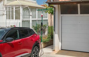 Ein rotes Auto vor einem Haus mit einer weißen Garagentür, einem grünen Gartenschlauch und einem Zaun aus Maschendraht.