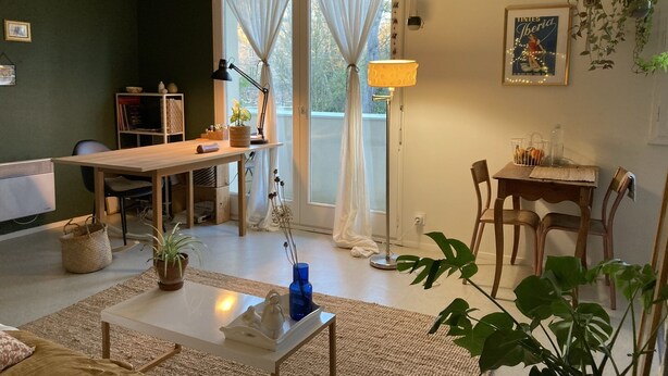 Een gezellige woonkamer met een houten tafel, een witte salontafel en een bank. Er is een boekenplank, een lamp en een plant in pot.