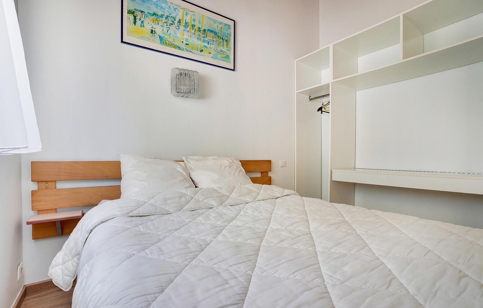 Ein Schlafzimmer mit einem Bett, einem Holz-Kopfteil, einer weißen, gesteppten Bettdecke, einem gerahmten Gemälde an der Wand und einem weißen Kleiderschrank mit geöffneten Türen.