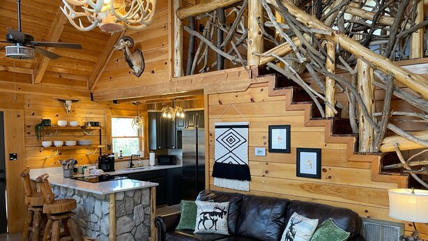 A rustic cabin interior with a stone fireplace, wooden furniture, a black leather couch, and a staircase made of logs. - Wildlife Cabin - By Magic Springs, Off-Road Park (Hot Springs)