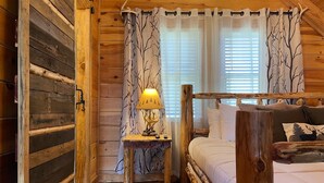 A wooden cabin bedroom with a bed, a nightstand with a lamp, and curtains with a tree pattern. - Wildlife Cabin - By Magic Springs, Off-Road Park (Hot Springs)