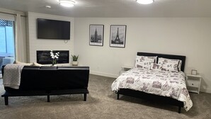 Une chambre avec un canapé noir, un lit recouvert d’une couvre-lit à motifs, deux tableaux encadrés représentant la Tour Eiffel, un téléviseur à écran plat fixé au mur et une petite table de chevet.