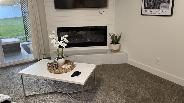 Un salon moderne avec un téléviseur à écran plat fixé au mur, une cheminée, une table basse ornée d’un vase de fleurs d’orchidées blanches et un tableau encadré représentant l’Empire State Building.