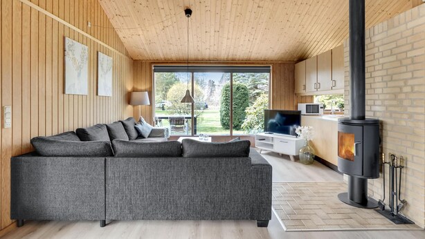 A modern living room with a grey sectional sofa, a TV, a fireplace, and a large window offering a view of the outdoors. - Awesome home in Væggerløse with kitchen (Væggerløse)