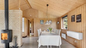 A cozy room with a wood-burning stove, a dining table with chairs, a hanging light fixture, and a window with a view of greenery. - Awesome home in Væggerløse with kitchen (Væggerløse)